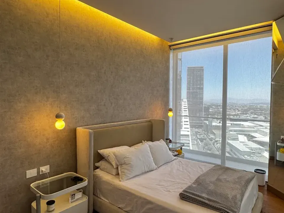 Recámara con cama de cabecera tapizada, piso de madera, luz cálida en el techo y ventanal con vista a la ciudad