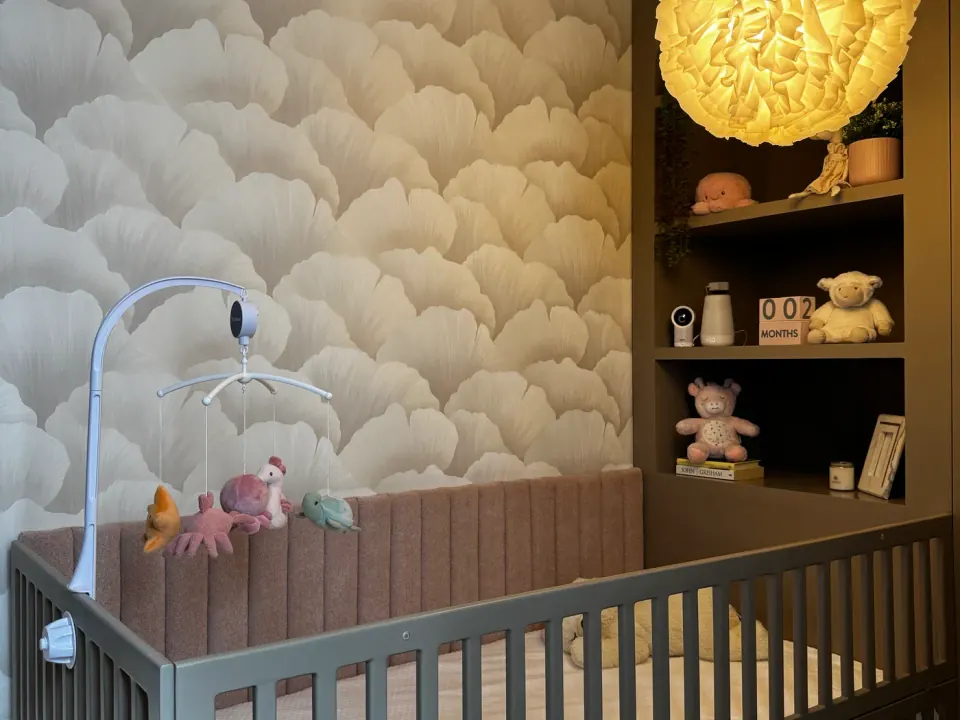 Cuna con móvil de peluches frente a papel tapiz blanco, con lámpara encendida y repisas en nicho a la derecha