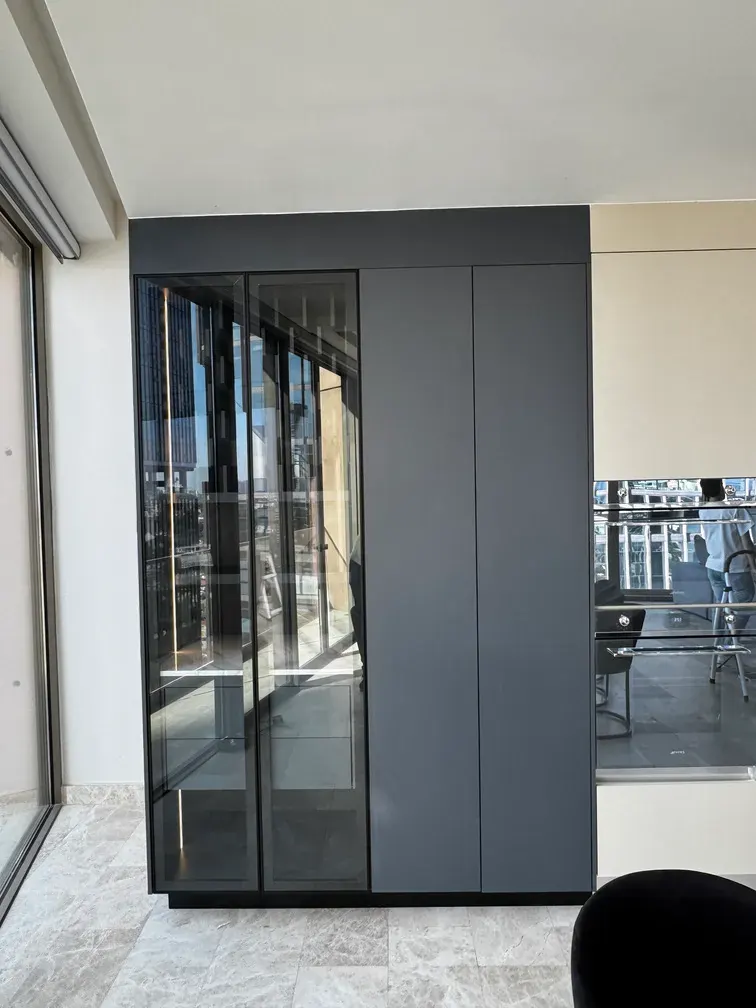 Mueble gris oscuro con puertas de vidrio ahumado y luz LED integrada, junto a la cocina y su piso claro veteado