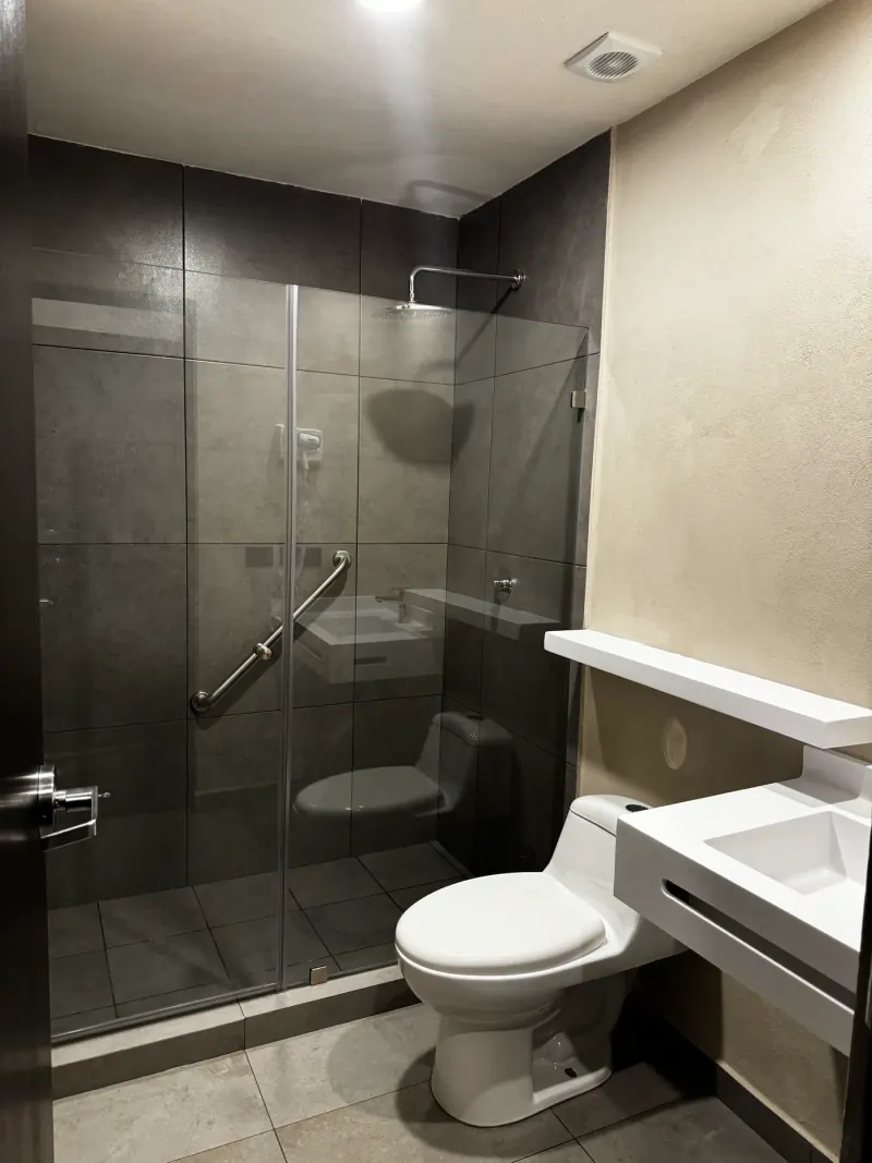 Baño con regadera cerrada con cristal, revestimiento gris de formato grande, barra de apoyo y lavabo blanco