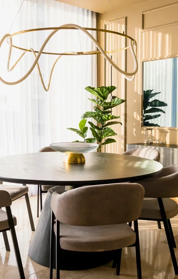Comedor de mesa redonda con sillas tapizadas, lámpara de aro dorado y espejo junto a ventanales con cortinas ligeras