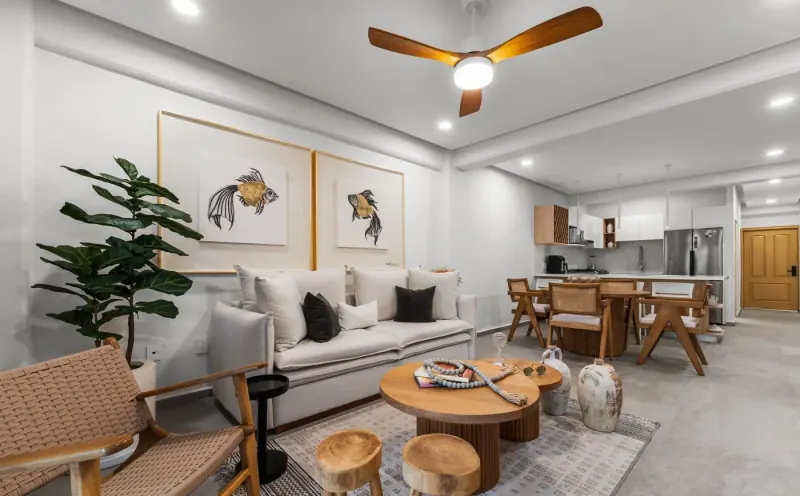 Sala con ventilador de aspas color madera, planta grande, sillón tejido y comedor de madera junto a la cocina