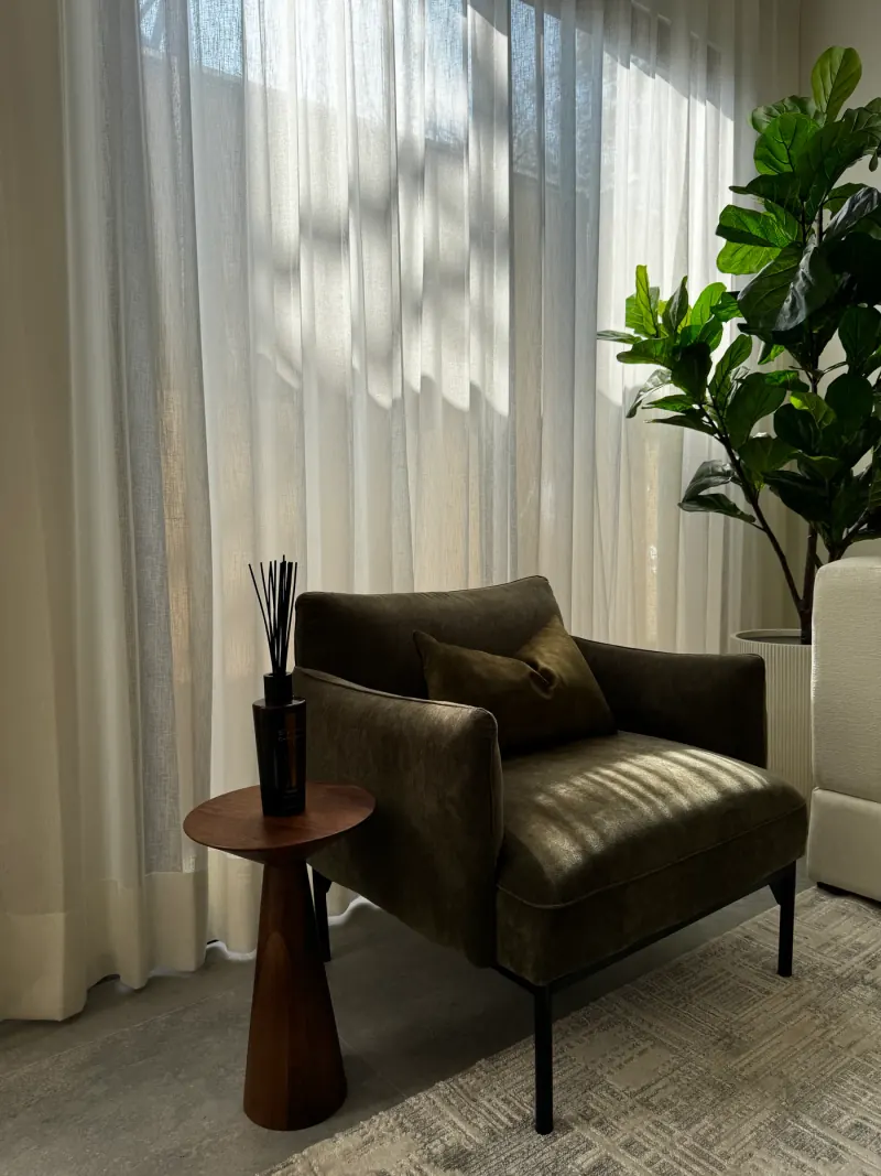 Rincón con sillón verde oliva, mesa auxiliar de madera, difusor de aromas y cortina ligera junto a una planta grande