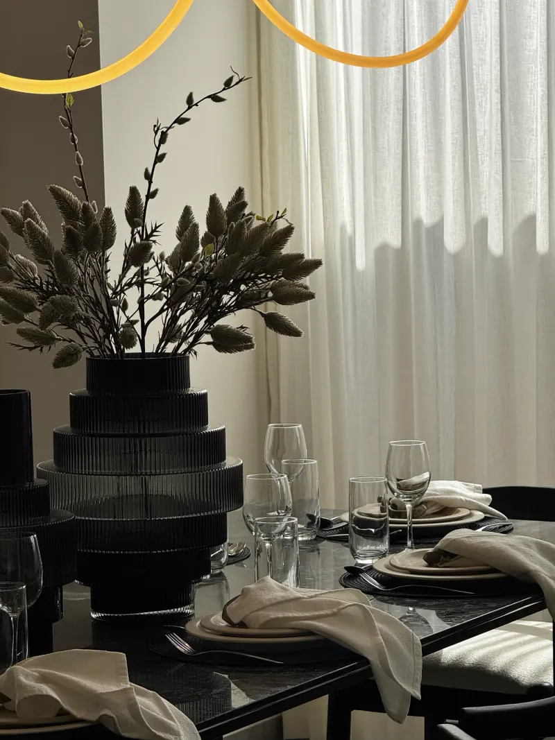 Detalle de mesa servida: florero acanalado oscuro con ramas, copas, platos beige y servilletas sobre cubierta negra