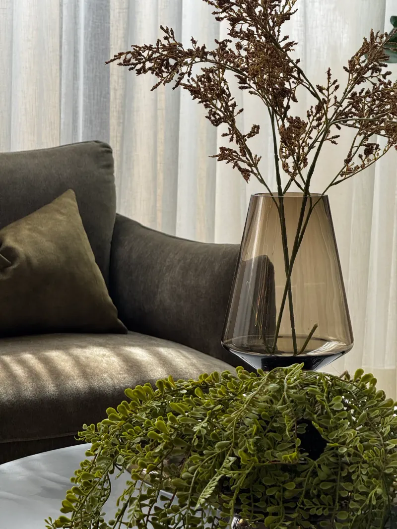Detalle de sala: florero de cristal ahumado con ramas, planta colgante y sillón oliva con cojín del mismo tono