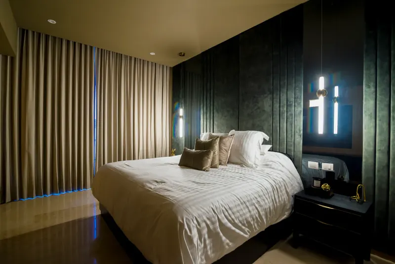 Cama con cojines beige junto a cortinas gruesas, paneles verdes tapizados y luminarias verticales colgantes