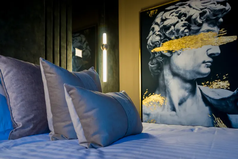 Detalle de cojines grises sobre ropa de cama blanca, con un cuadro de busto clásico y trazo dorado al fondo, Zapopan